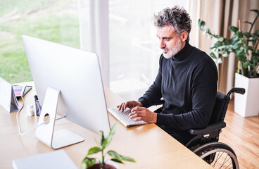 Handsome man in wheelchair working from home office, using computer in office. Telecommuting and home office.