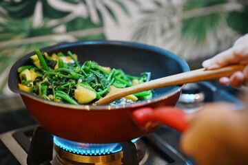 Person stirring fresh vegetables in a red pan on a stove, with a lush green background, depicting home cooking and healthy eating.