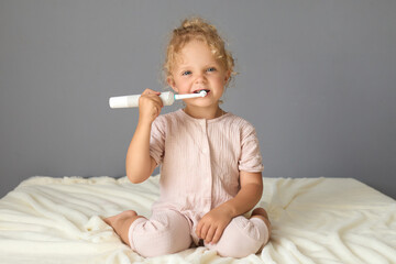 Cute Caucasian female child engaging in her dental care routine using toothpaste and toothbrush to clean her teeth while sitting on the bed in the bedroom