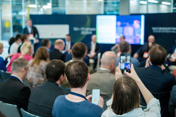 Business professionals attend a conference, engaging with the speaker and capturing moments on their phones.