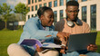 © Yuliia - Two African American friends students high school pupils classmates university college academy in park outdoors learning studying together laptop computer online e-learning laughing drinking coffee