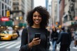 © Khawla - A Black Pretty Woman with radiant smile and holding her phone while walking down the street, with blurred city buildings.