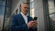 © Yuliia - Caucasian senior mature male employer leader boss retired old middle-aged businessman business entrepreneur walking in city going to office outdoors looking in smartphone typing chatting mobile phone