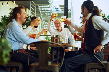 Business professional, smiling men in formal wear meeting at outdoor bar and raising glasses of beer in celebratory toast. Cheers to successful partnership. Concept of business, Oktoberfest, team