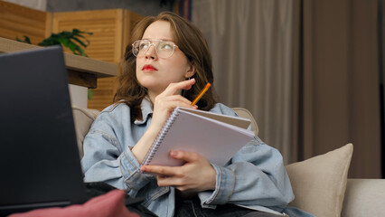 Seated on a sofa, a woman in casual attire with glasses writes in a notebook, her gaze fixed on a laptop screen. This photo emphasizes the dedication and focus required for successful remote work or