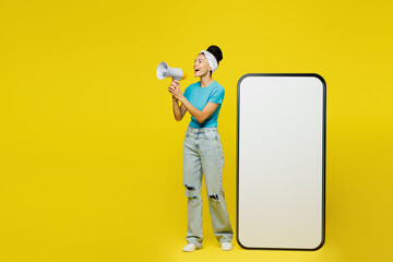 Wall Mural - Full body young happy Latin woman wears blue top t-shirt headband casual clothes big huge blank screen mobile cell phone smartphone with area scream in megaphone isolated on plain yellow background.