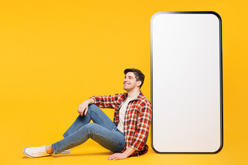 Wall Mural - Full body side view young man he wears red checkered shirt casual clothes sit near big huge blank screen mobile cell phone smartphone with area isolated on plain yellow background. Lifestyle concept.