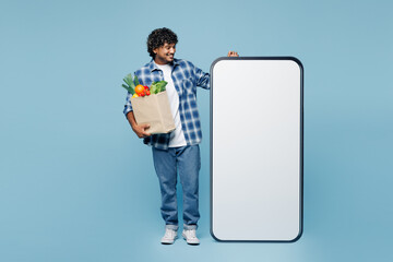 Wall Mural - Full body young Indian man wear shirt hold craft bag for takeaway with food products stand look at big huge blank screen mobile cell phone isolated on plain blue background Delivery service from shop