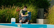 © StockMediaSeller - Pool technician using a tablet by the pool, surrounded by greenery and a chemical container, focused on maintenance tasks.