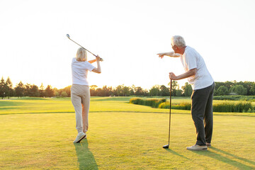 elderly senior couple in uniform playing golf on golf course at sunset, old man instructor teaching woman how to hold golf club and swing ball, grandfather helping grandmother play outdoors