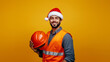 © Favio - 20s Hispanic man, construction worker wearing a santa hat and holding a helmet isolated on yellow background.