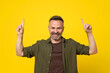 © ReyRomMedia - Handsome middle age hoary stubble senior man dressed in green shirt over isolated yellow background showing and pointing up with finger by two hands number one while smiling confident, excited, happy.