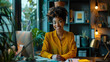 © Jirawatfoto - A smiling woman in yellow shirt sits at desk surrounded by plants and cozy workspace.