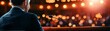 © ธนากร บัวพรหม - Speaker in formal attire at a podium addresses audience with bright lights and bokeh effect in background.