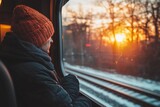 Fototapeta  - A contemplative man wearing a warm knitted hat gazes out the window of a train as the sun sets, casting a warm glow over the snowy winter landscape.