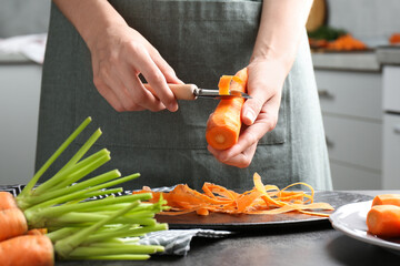 Poster - Woman peeling fresh carrot at dark gray table in kitchen, closeup