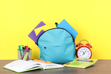 Poster - Backpack with different school stationery and alarm clock on wooden table against yellow background