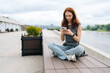 © dikushin - Relaxed pretty woman with long red hair sitting by river, using smartphone smiling looking to screen, enjoying break from modern life in nature. Calm redhead female happily using phone by river.