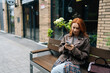© dikushin - Pretty young woman in trendy leather jacket sitting on bench outdoors, happily browsing smartphone. Stylish redhead female using mobile phone on city bench, enjoying free time in European city.