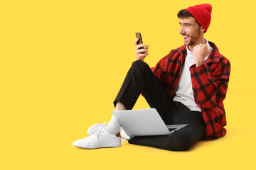 Wall Mural - Happy young man with laptop using mobile phone on yellow background