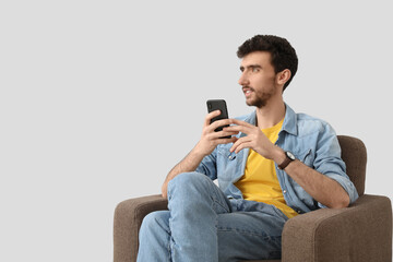 Wall Mural - Handsome young man using mobile phone in armchair on light background