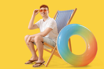 Wall Mural - Young man with inflatable ring resting in deck chair on yellow background