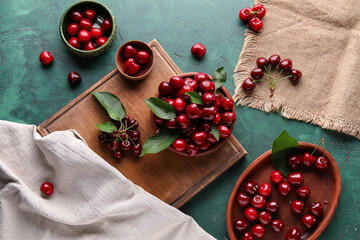 Bowls and board with sweet cherries on green background