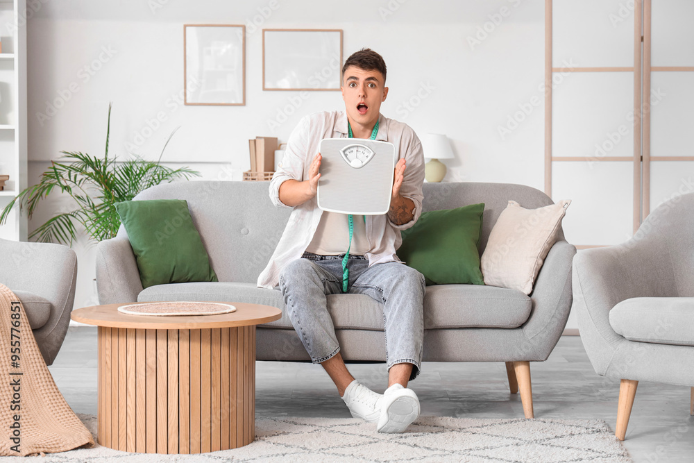 Shocked young man with weight scales sitting on sofa at home