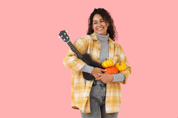 Poster - Beautiful young happy African-American woman with pumpkins and ukulele on pink background