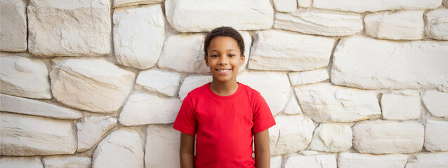  A Black boy wearing a red T-shirt and laughing with his back against a stone wall