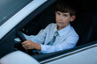 © Emvats - A young boy in a white shirt and blue plaid tie sits in the driver's seat of a car, holding the steering wheel with a serious expression. The image captures a moment of curiosity and playfulness