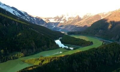 Sticker - The camera flies over a lush green valley surrounded by towering snow-capped mountains. A winding river snakes through the landscape, reflecting the golden rays of the setting sun.