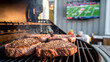 © ontsunan - Grilling steaks on a barbecue grill outdoors with a TV showing a football game in the background, perfect for a backyard party.