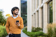 © Liubomir - Hispanic businessman in orange sweater holding phone while walking outside office building. Smiling confidently and looking forward, showcasing modern urban work environment , professional lifestyle.