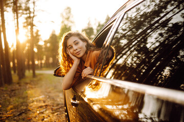 Wall Mural - A woman  joyfully leans out of a car window, basking in the golden light filtering through the tall trees during sunset in a tranquil forest setting. The concept of active lifestyle, travel, tourism, 