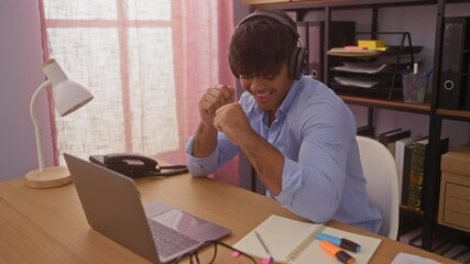 Wall Mural - Young hispanic man listening to music with headphones in an office setting, working at a wooden desk with a lamp, notebook, markers, and a laptop, enjoying a moment of relaxation.