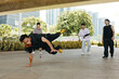 © DragonImages - Group of breakdancers showcasing energetic skills on urban pavement, surrounded by green foliage and tall buildings in background. Performers display athleticism and coordination