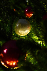 Golden glittery ball hanging on green plastic Christmas tree in darkness, lit by lights of garlands. Vertical photo, close-up, copy space. Template, background for greeting card.