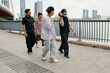 © DragonImages - Group of diverse friends strolling along waterfront promenade with modern buildings in background, some wearing casual attire, engaging in lively conversation