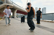 © DragonImages - Three young men dancing on an urban sidewalk with a bridge and cityscape in background One is performing in front while two others watch, all engaged in freestyle dancing activity