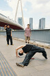 © DragonImages - Group of young men performing breakdance moves on urban bridge with city skyline in background, showcasing dynamic movement and athleticism