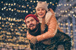 © deagreez - Photo of positive cheerful couple guy hug piggyback girlfriend look x-mas christmas evening lights outdoors wear scarf coat headwear
