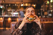 © zvkate - Close-up portrait of a woman in a fast food restaurant eating a hamburger. Hungry woman eating a burger sitting in cafe. Girl bite bbq burger, look joyful. Cheat meal day concept.