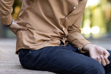 Individual seated on bench outdoors showing signs of discomfort due to lower back pain. Casual attire, hand on back indicating ache or injury. Expresses physical discomfort, health concern