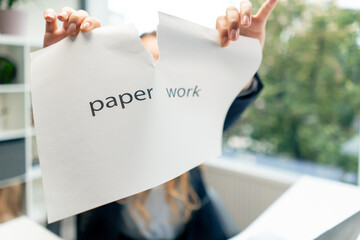 close-up in a modern office with a large window a beautiful female worker in a blue shirt sitting at a table directly at the camera tears a white sheet of paper with the inscription paperwork