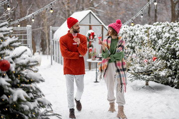 Man and woman talk and drink while spending winter time together at beautifully decorated snowy backyard. Young family celebrating Near Year's holidays