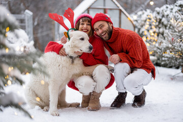 Lovely couple hug with their cute dog wearing toy deers horns at snowy backyard. Young family spending happy winter time together outdoors