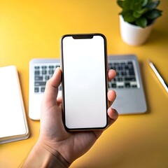 A close up shot of a hand holding a smartphone with a blank white screen against a yellow background with a laptop in the background.