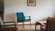 © BISO - A mid-century style living room with a white wall and an art poster frame. There is a wooden chair with a teal cushion near the wall and a wooden floor.