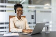 © Liubomir - Confident African American businesswoman using laptop and headset for customer support. She is smiling, focused, and working efficiently in modern office setting.